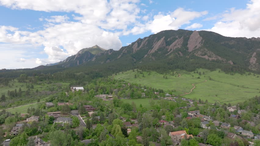 Wide pan right aerial view of warm sun hitting Boulder Colorado Flatiron mountains above Chautauqua Park with full green pine trees and blue skies with clouds on a beautiful summer day for hiking