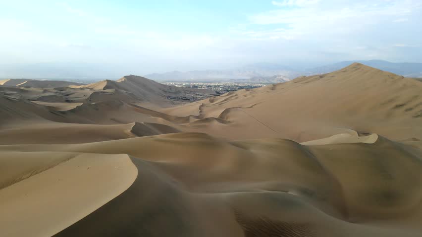 Aerial shot drone flies backwards over sand dunes near desert oasis Huacachina, Peru with Ica in distance