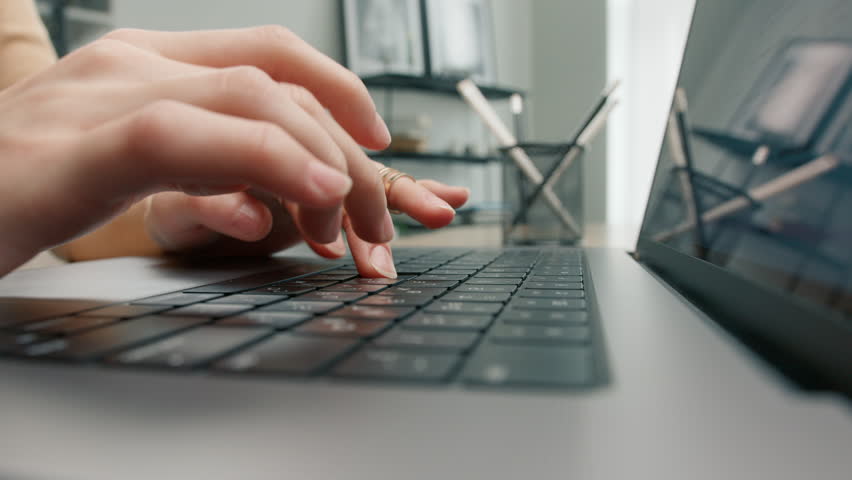 Concept of performing online work project, slow motion. Close up of unrecognizable female hands with slim fingers working on laptop computer at desk in modern office room on background of large window
