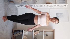 Vertical video. Jumping Jack. Home exercise. Happy young energetic fit woman in sportswear training in light room interior. Spread feet and hands overhead.