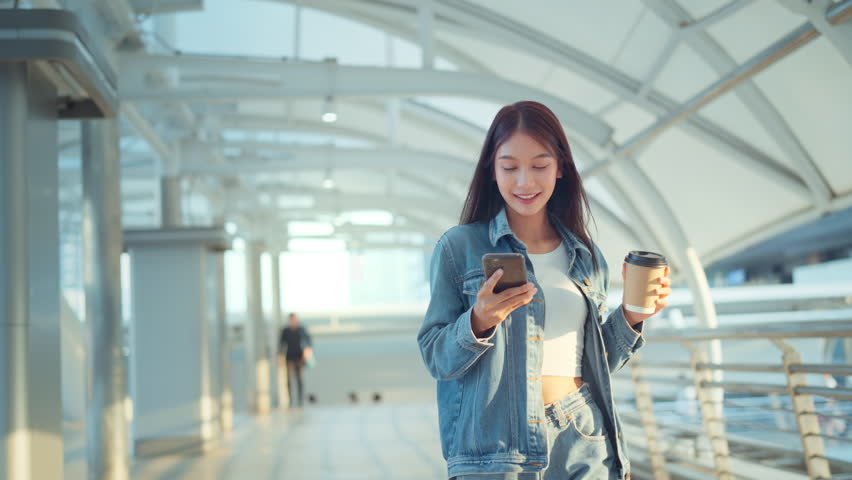 Young asian woman walking with mobile phone and coffee cup in city, Cheerful woman relaxing with cellphone and hot drink outdoors. Royalty-Free Stock Footage #1105267159