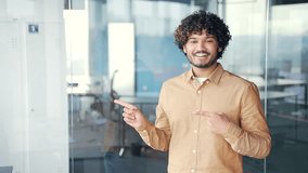 Young smiling man pointing finger at empty space for writing text while standing in modern office. Positive businessman looking at camera. Template for advertising. Copy space for goods promotion - Powered by Shutterstock - Get 15% off with code: PIKWIZARD15