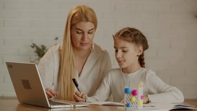 Young mother helps her daughter with homework on her laptop. Cute mom and daughter do their homework.