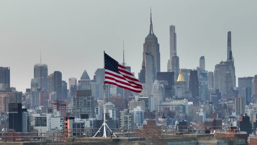 Brooklyn , NY , United States - 06 13 2023: American flag waving in front of Empire State Building and midtown Manhattan skyline. Long aerial zoom of Brooklyn Bridge USA flag with New York City skylin