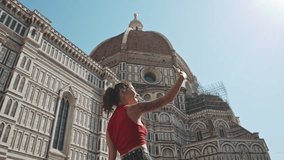 Rear view of young asian girl taking selfie on smartphone or mobile phone in front of dome of cathedral. Woman traveling on foot in Italy. Summer cityscape in Florence. Lifestyle and outside concept.