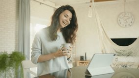 Attractive brunette communicates via video link on tablet sitting at table, drinking tea. Young woman laughs, gesticulates a lot, tosses her curly hair. High quality 4k footage