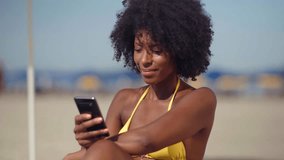 Cheerful african american young man hold mobile phone using app, social network, video call, watching funny videos and laughing sitting at the beach lifestyle. Concept online browsing film.
