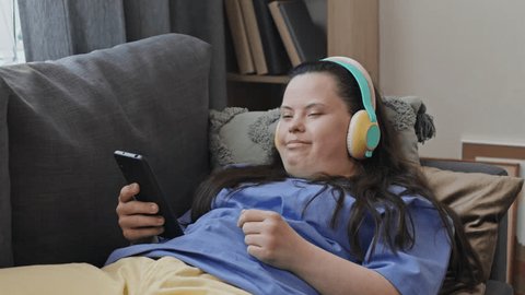 Medium shot of cheerful young Caucasian girl with Down syndrome wearing headphones relaxing in living room enjoying music Video Stok