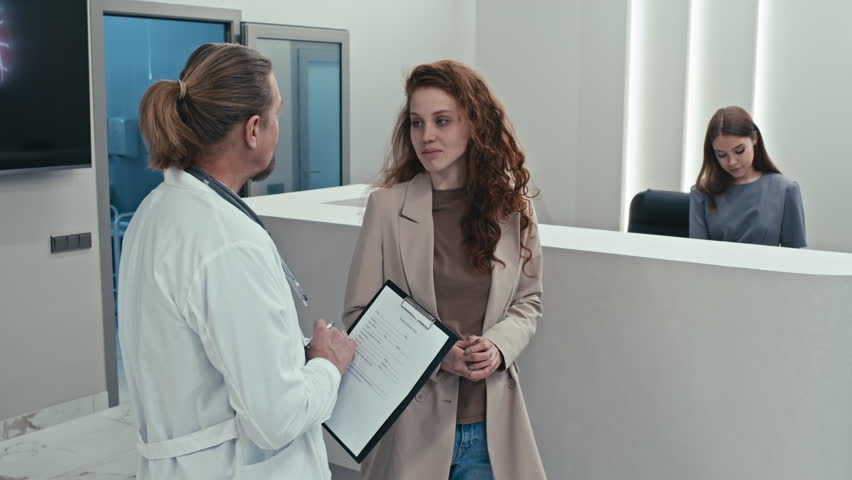 Mature male doctor and young female patient standing in modern hospital lobby having conversation