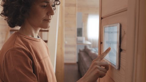 Selective focus shot of Caucasian woman living in modern wooden house equipped with home automation system changing indoor temperature using tablet on wall Video de stock