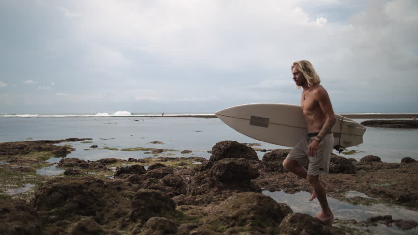 Male surfer walks along sharp rocks on beach to sea or ocean to ride surfboard. Sporty Caucasian male surfer on seacoast. Concept of skills development, development and expansion of own abilities