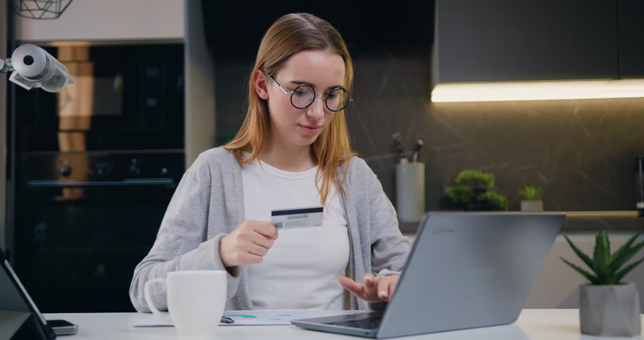 Cheerful girl uses laptop and holding credit card in the hands, makes purchases in the online store at home. Internet sale,finance, ecommerce payment online shopping and banking on internet typing