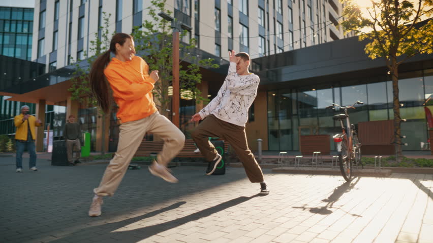 Two Professional Hip Hop Dancers Jamming On City Street Among Modern Buildings. Male And Female Newstyle Performers Freestyling, Fashionable Friends Supporting and Livestreaming Via Smartphone.