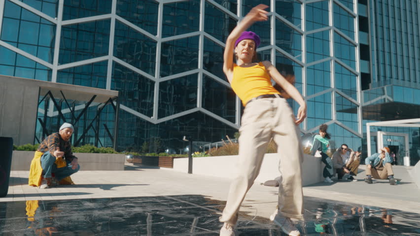 Young And Stylish Female Hip-Hop Dancer Improvising On City Street. Fashionable Group Of Friends Relaxing On Background, Supporting Professional Street Style Performer Practising Choreography.