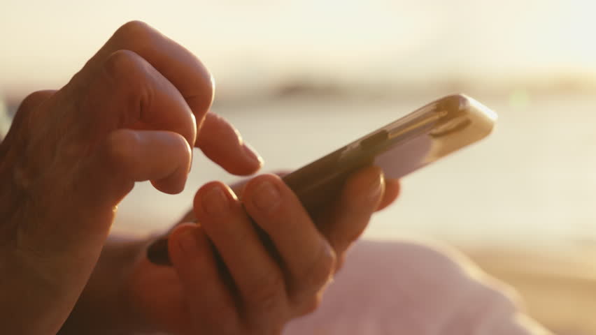 Close-up macro footage of aged female hands holding cell phone scrolling and texting message from social media against golden light on sunset at seaside. Elderly lady tapping screen of mobile phone.  Royalty-Free Stock Footage #1105322351