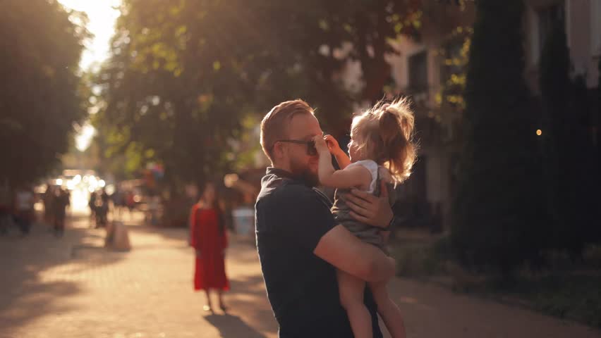 A father walks with his daughter in the park. Stylish bearded father holds little two year old daughter in his arms, daughter takes off his sunglasses and has fun laughing at sunset