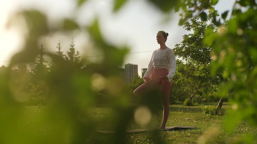 Tracking shot of young sporty woman practicing yoga standing in Vrksasana exercise, Tree pose on yoga mat in park in evening during sunset. Beautiful sunset sky and landscape on background.