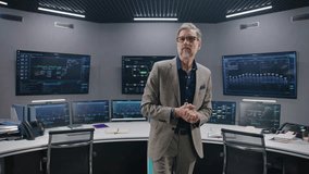 Mature supervisor in business suit greets programmers in monitoring room and looks at camera. Big data engineers start work. Multiple big screens with displayed real-time analysis charts. Dolly shot. - Powered by Shutterstock - Get 15% off with code: PIKWIZARD15