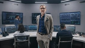 Mature supervisor in business suit greets IT engineers in monitoring room and looks at camera. Big data engineers start work. Multiple big screens with displayed real-time analysis charts. Dolly shot. - Powered by Shutterstock - Get 15% off with code: PIKWIZARD15