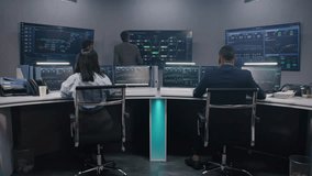 Mature big data scientist greets IT engineers in monitoring room. Support specialists sit at computers and control real-time analysis charts. Multiple big screens with blockchain network. Dolly shot. - Powered by Shutterstock - Get 15% off with code: PIKWIZARD15
