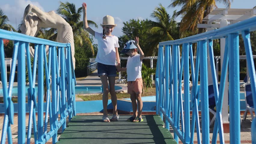 Children have fun on vacation at the hotel.Brother and sister jump and wave their arm.Summer holidays for children.A boy and a girl,holding hands,stand on a bridge against the background of a fountain
