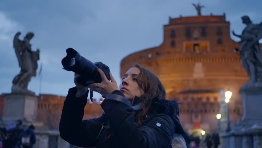 Young Latina woman in warm jacket taking photos on professional photo camera, shooting architecture of Ponte Sant Angelo bridge with lanterns in evening, Rome, Italy