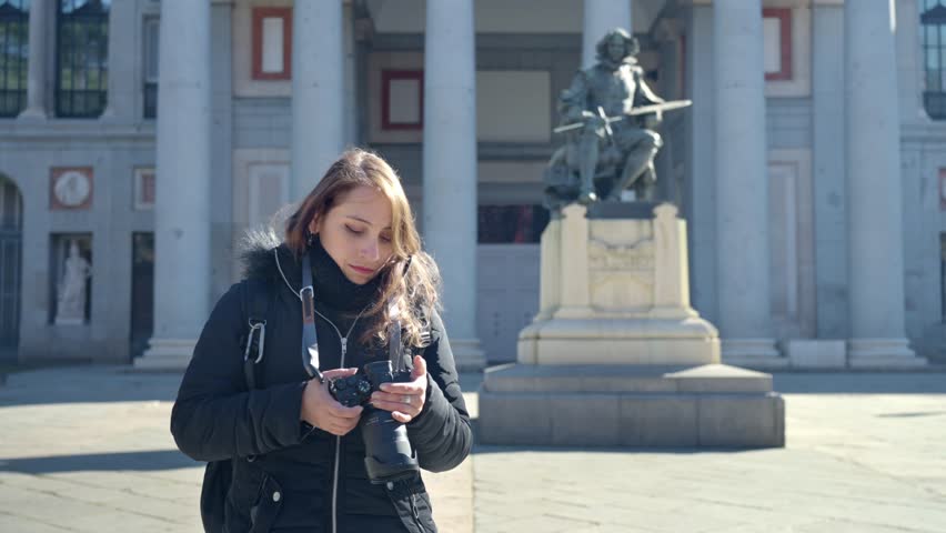 Smiling young Latin woman in warm clothes with professional photo camera looking at camera while standing near sculpture at Prado Museum in Madrid, Spain