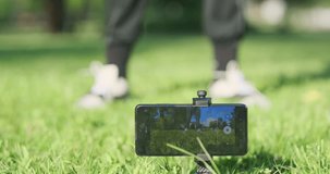 Fitness trainer records himself video on smartphone, working out in park. Squats on green grass. Online fitness.