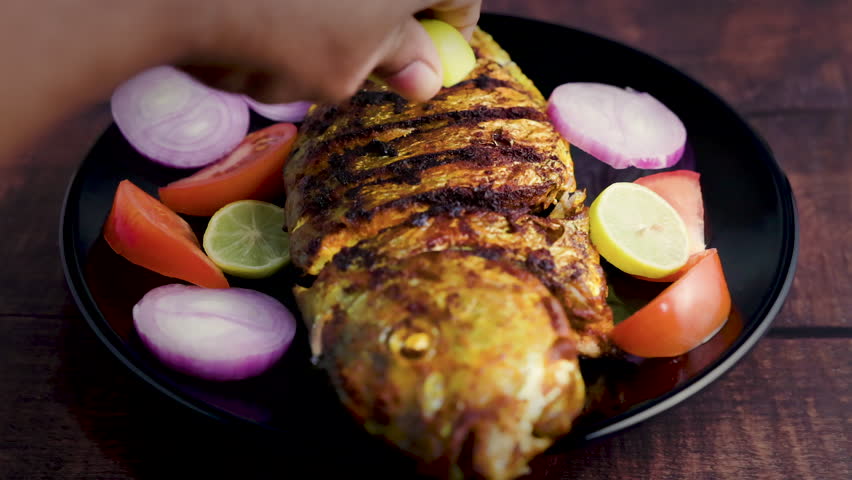 Delicious fried fish or fish fry on a black plate with onion, tomato, and lemon. Spicy Indian Kerala style fish fry on a wooden table. Lemon squeezed over grilled fish. | Shutterstock HD Video #1105343563