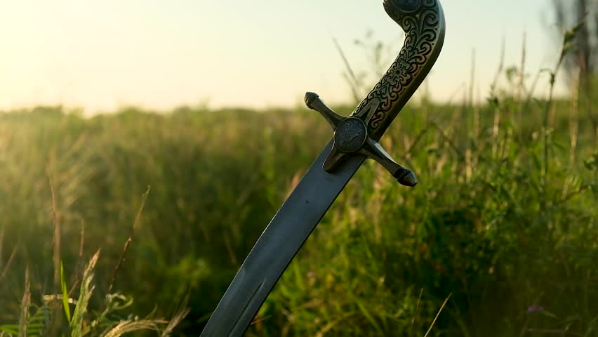 The saber of the Ukrainian Cossacks is stuck in the ground, among the grass, against the background of the sunset. Beautiful bokeh appears in the frame. The concept of freedom. Close-up.