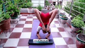 Camera zooms in on a young Indian girl doing yoga playing a keyboard with both hands and putting her feet in the air while doing yoga