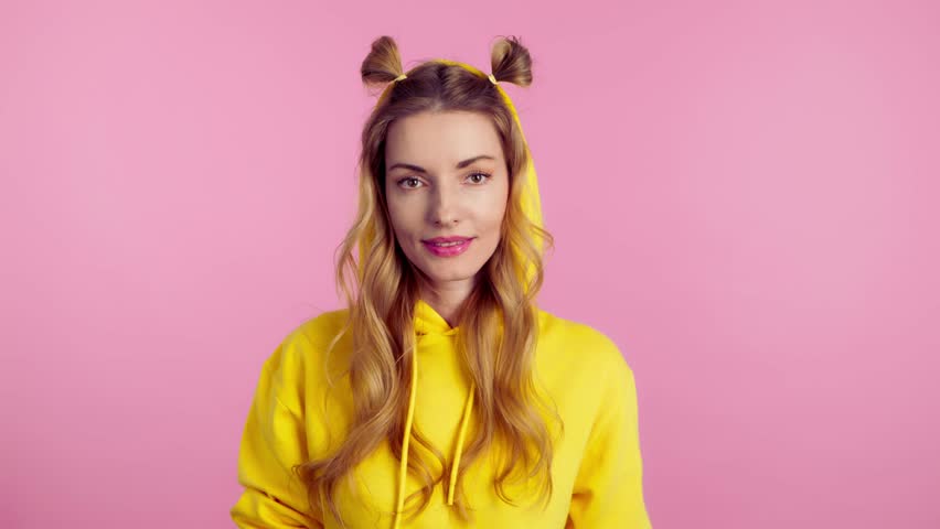 Woman sends air kiss smiling looking at camera. Happy positive emotional blonde girl. Joyful cheerful pretty lady in yellow hoodie, smile face. Indoors studio, isolated on pink background | Shutterstock HD Video #1105356559
