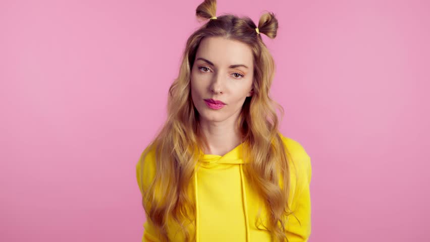 Woman sends air kiss smiling looking at camera. Happy positive emotional blonde girl. Joyful cheerful pretty lady in yellow hoodie, smile face. Indoors studio, isolated on pink background | Shutterstock HD Video #1105356575