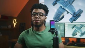 African American 3D designer speaks on camera using microphone, records video tutorial about 3D modeling and animation. 3D aircraft design and software interface on computer and big digital screen.