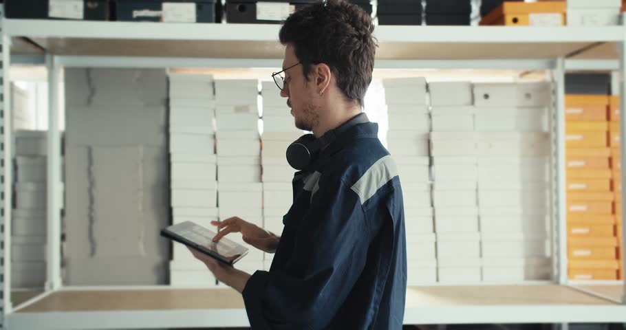 A man in a blue uniform with a tablet in his hands is preparing for stocktaking in a warehouse with yellow and white boxes and puts on glasses