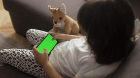 The girl is sitting on the couch and playing games on her phone. The phone is in focus, with a chrome key. eside the girl, there is a plush toy kangaroo sitting and looking at the screen.