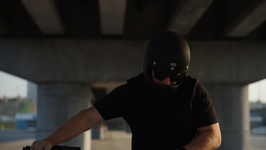 A biker holds the motorcycle by the handlebars and walks on the asphalt with the bike muffled. The biker has a black helmet with glasses. A serious biker on a motorcycle walks under a road bridge. A b