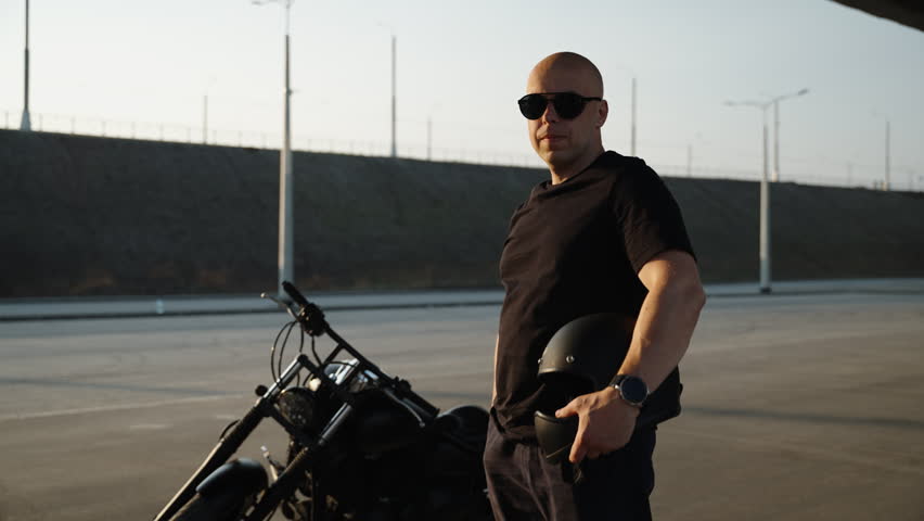 A man stands next to a black chrome motorcycle and looks into the camera. Black T-shirt and pants, along with biker sunglasses. The motorcycle stands in the summer parking lot under the bridge. Royalty-Free Stock Footage #1105380117