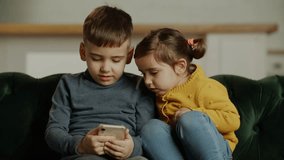 Siblings playing game on modern phone while sitting together on comfortable sofa. Children playing virtual game on smartphone, watching online funny content on internet