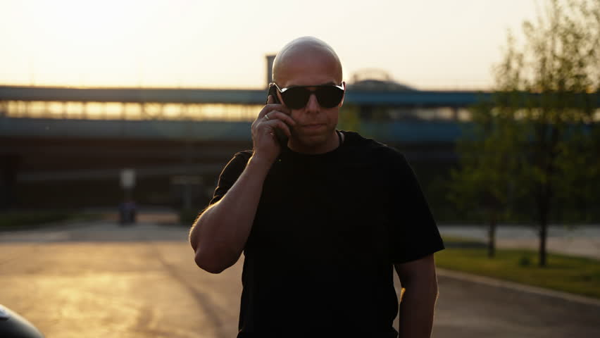 A brutal man in black clothes and sunglasses walks down the street and listens to a voice message on his phone. Talking on the phone. Communication between people. Sunset lighting behind a man in blac