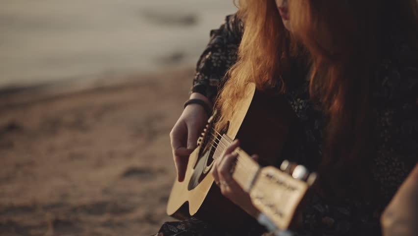 Red-haired hipster girl in beautiful sunglasses plays the guitar. Pretty female sings a song on sunset background. Vacation at the sea. Atmosphere of love. Self isolation, travel concept.