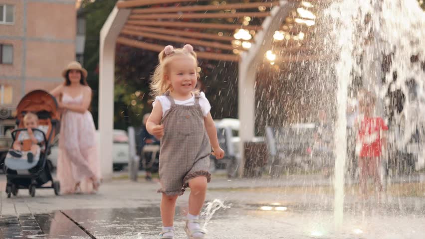 Happy little girl is playing with the stream of water, running on the water. A child is playing near a fountain. Fountain in the town square outdoors. Summer vacation and rest Royalty-Free Stock Footage #1105399173