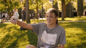 Portrait of a teenager girl in glasses in the park with a phone, she sits on the green grass and takes a selfie. 