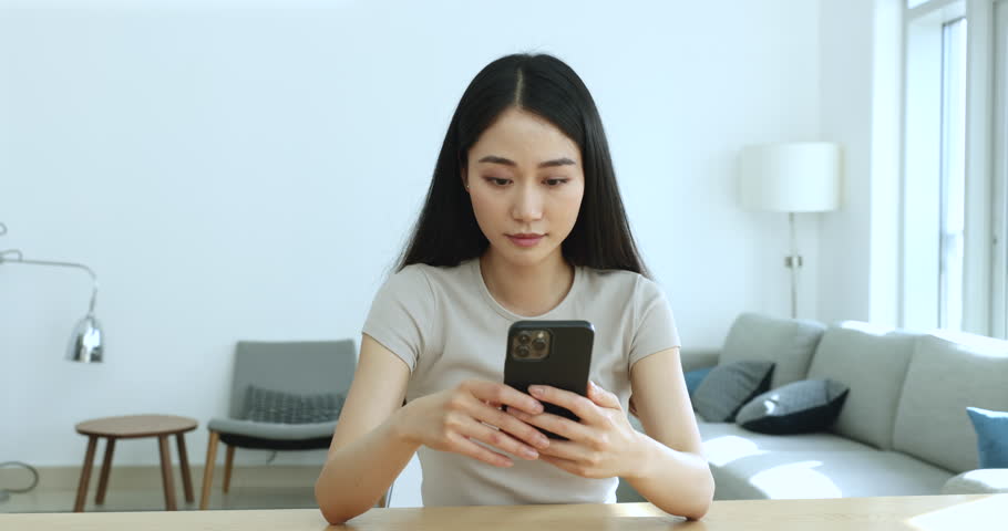 Attractive young 25s Asian female sit at desk looks at smart phone screen and frowning while read unpleasant message, get bad news, experiences negative emotions due to problems with cellphone usage Royalty-Free Stock Footage #1105408813
