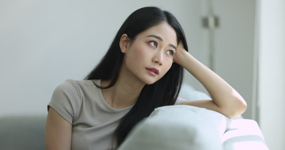 Sad Asian beautiful woman sits on couch deep in unhappy thoughts, looking away leaning head on hand, have melancholic mood, thinks on quarrel with beloved man, feel boredom on weekend alone at home Royalty-Free Stock Footage #1105412217