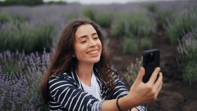 Young freelancer girl brunette woman student sitting on lavender field holding mobile phone gadget relaxing outdoors chatting online with friend video call talking on smartphone webcam using virtual n