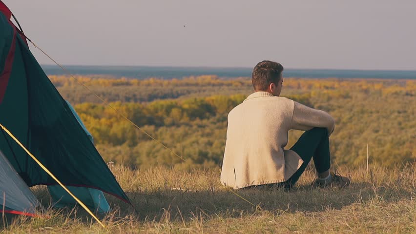 lonely guy in brown pullover sits by blue tent shadow and admires pictorial autumn forests slow motion backside view