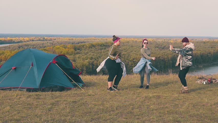 joyful young women dance at tent in touristic camp on steep river bank against pictorial landscape in autumn slow motion