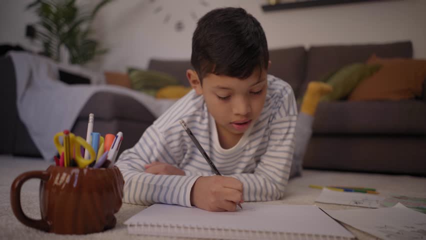 Little Latino boy draws quietly lying in living room at home. Concentrated child doing school homework at weekend. Concept of kids activities on vacations and family free time in childhood.  Royalty-Free Stock Footage #1105425565