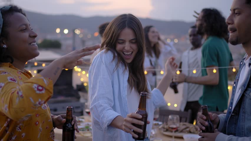 Three young excited multi-ethnic friends dance at rooftop barbecue party. Cheerful people jump and toast with beer celebrating BBQ in the evening. Generation z students have fun on summer vacation. Royalty-Free Stock Footage #1105425581